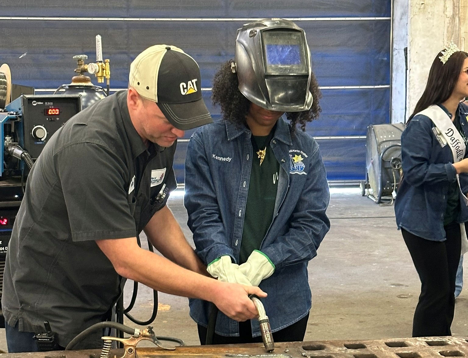 A man helps a student in welding gear at a work station. 